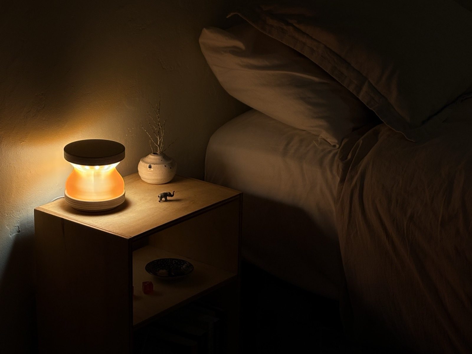Apollo lamp with a warm amber hourglass shade and Baltic-birch cap and base, glowing on a wooden bedside table next to a made bed at night