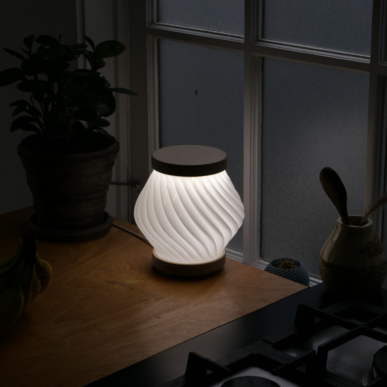 Apollo lamp with a white spiral-fluted shade glowing on a kitchen counter at dusk, its power cable trailing behind it